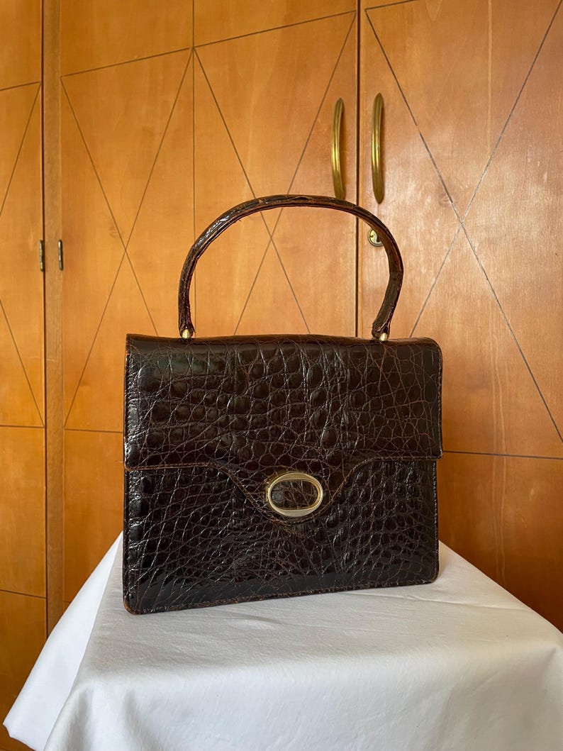 Puede incluir: Un bolso vintage marr&oacute;n con un patr&oacute;n texturizado similar al de un cocodrilo. El bolso tiene un asa curva, un cierre ovalado dorado y detalles dorados. El bolso est&aacute; sobre una superficie blanca.