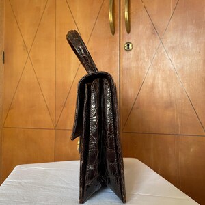 Puede incluir: Un bolso vintage marr&oacute;n oscuro con un patr&oacute;n texturizado similar al de un cocodrilo. El bolso tiene un asa superior, una forma rectangular y un cierre dorado. El bolso est&aacute; sobre una superficie blanca.