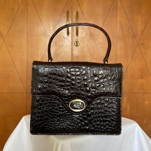 Puede incluir: Un bolso estructurado de color marr&oacute;n oscuro con textura de cocodrilo. El bolso tiene un asa superior, un cierre dorado y forma rectangular. El bolso est&aacute; sobre una superficie blanca.