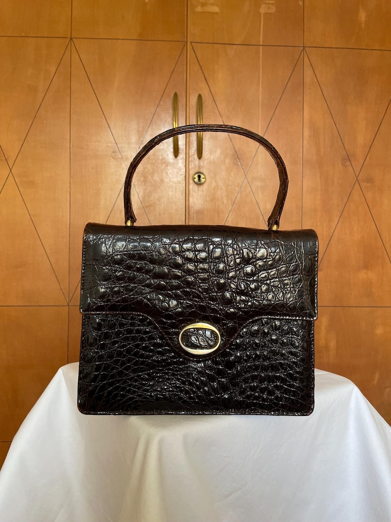 Puede incluir: Un bolso estructurado de color marr&oacute;n oscuro con textura de cocodrilo. El bolso tiene un asa superior, un cierre dorado y forma rectangular. El bolso est&aacute; sobre una superficie blanca.