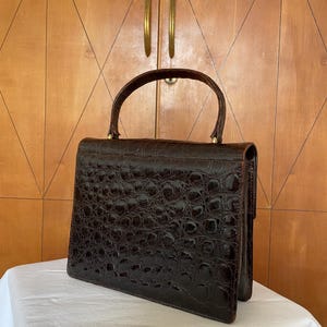 Puede incluir: Un bolso vintage marr&oacute;n oscuro con un patr&oacute;n texturizado similar al de un cocodrilo. El bolso tiene una forma estructurada, un asa superior y un cierre de broche. El bolso est&aacute; sobre una superficie blanca, con un armario de madera en el fondo.
