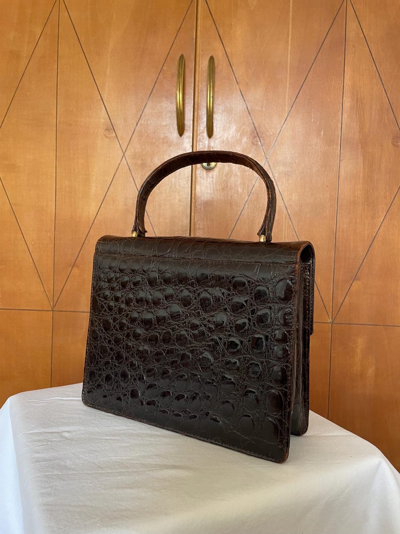Puede incluir: Un bolso vintage marr&oacute;n oscuro con un patr&oacute;n texturizado similar al de un cocodrilo. El bolso tiene una forma estructurada, un asa superior y un cierre de broche. El bolso est&aacute; sobre una superficie blanca, con un armario de madera en el fondo.