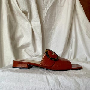 May include: A pair of brown leather slide sandals with a tortoiseshell buckle. The sandals have a low heel and a square toe design. The footwear is displayed on a white surface.