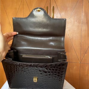 Puede incluir: Bolso de mano marr&oacute;n con textura de cocodrilo, abierto. Cuenta con un cierre y herrajes dorados. El interior revela un bolsillo con cremallera y una solapa. El bolso est&aacute; abierto, mostrando su interior.