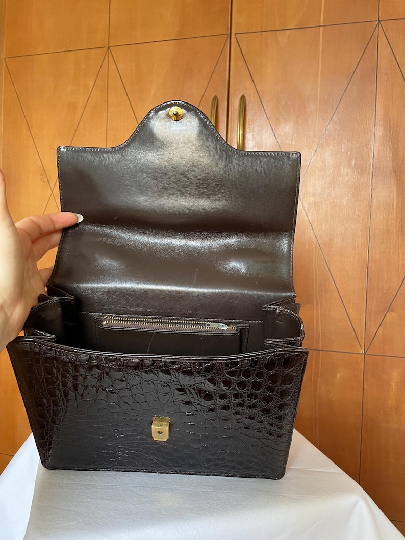 Puede incluir: Bolso de mano marr&oacute;n con textura de cocodrilo, abierto. Cuenta con un cierre y herrajes dorados. El interior revela un bolsillo con cremallera y una solapa. El bolso est&aacute; abierto, mostrando su interior.
