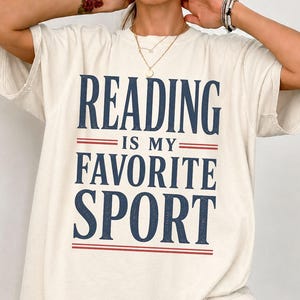 May include: An off-white short-sleeve t-shirt featuring the dark blue text "READING IS MY FAVORITE SPORT" printed on the front. The design includes two horizontal red lines above and below the phrase "IS MY" in a clean, centered layout.
