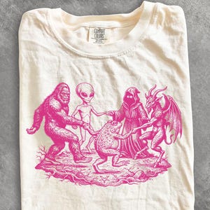 Op de afbeelding: Een crèmekleurig t-shirt met een felroze grafische print van een Bigfoot, een alien, een figuur met een kap, een pad en een gevleugeld geitachtig wezen die elkaars hand vasthouden in een cirkel op een stuk grond met een pentagram.