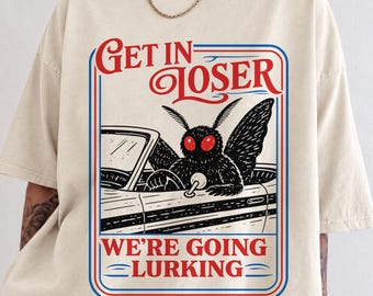 May include: A cream-colored short-sleeve t-shirt featuring a graphic print of a black moth with red eyes driving a convertible car. The text reads GET IN LOSER WE'RE GOING LURKING in red and blue lettering, enclosed within a rectangular border.