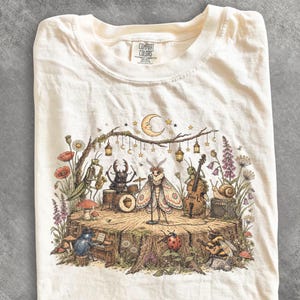 Op de afbeelding: Een crèmekleurig t-shirt met een grafische print van diverse insecten die muziekinstrumenten bespelen op een podium van een boomstronk. De illustratie toont een zingende mot, een kever op drums, een sprinkhaan op saxofoon en een bij met gitaar.