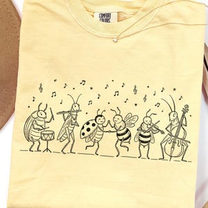 May include: A pale yellow t-shirt featuring a black line art illustration of a group of insects playing musical instruments, including a beetle on drums, a grasshopper on flute, a ladybug and bee dancing, and a bee playing a violin and cello.
