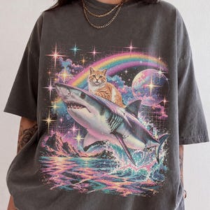 Könnte beinhalten: Ein anthrazitfarbenes T-Shirt mit einem Grafikdruck einer orangefarbenen getigerten Katze, die auf einem Hai reitet. Der Hintergrund zeigt einen bunten Regenbogen, einen leuchtenden Mond, funkelnde Sterne und eine Berglandschaft.