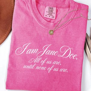 May include: A neon pink short-sleeved t-shirt featuring white cursive and serif text that reads, I am Jane Doe. All of us are, until none of us are. The shirt has a crew neckline and a Comfort Colors brand label visible at the inner collar.