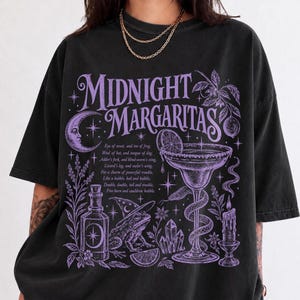Può includere: Una t-shirt nera a maniche corte con una stampa grafica viola e il testo Midnight Margaritas. Il design include una luna crescente, una rana con un cappello da strega, un bicchiere da cocktail con un serpente, una candela e una pozione.