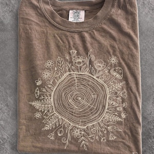 May include: A brown crew neck t-shirt featuring a white line art graphic of a tree stump surrounded by various flowers, ferns, mushrooms, and bees. The inner neck label displays the text Comfort Colors, 100% cotton, and made in Honduras.
