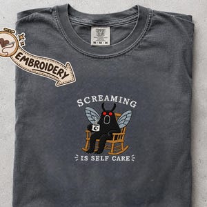 Può includere: Una t-shirt grigio antracite con un ricamo raffigurante una creatura nera simile a una falena con occhi rossi, seduta su una sedia a dondolo in legno mentre tiene una tazza. Il testo "SCREAMING IS SELF CARE" è scritto sopra e sotto.
