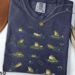 May include: A navy blue crew neck t-shirt featuring a repeating graphic print of various green frogs in different poses on lily pads and in water. The inner neck label reads Comfort Colors, Heavyweight Garment Dye, 100% Cotton, Made in Honduras, S P CH.