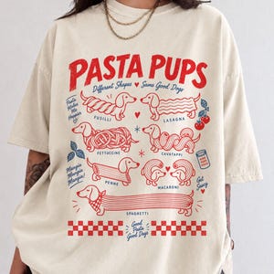 May include: A cream-colored t-shirt featuring a red and blue graphic print titled PASTA PUPS. The design displays various dachshund dogs illustrated as different pasta shapes including fusilli, lasagna, fettuccine, cavatappi, penne, macaroni, and spaghetti.