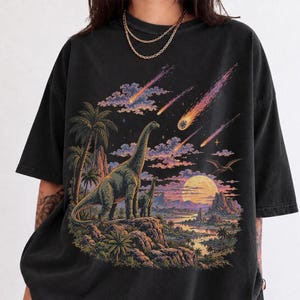 Op de afbeelding: Een zwart t-shirt met korte mouwen en een grafische print van twee langhalsdinosaurussen op een rotsachtige klif. De afbeelding bevat palmbomen, een grote gele maan, een rivier en meerdere feloranje en roze meteoren aan de hemel.