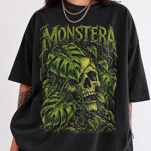 May include: A charcoal black short-sleeve t-shirt featuring a graphic print of a human skull intertwined with vibrant green monstera leaves and vines. The word MONSTERA is printed in a stylized, dripping green font at the top of the design.