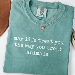 May include: A sage green short-sleeve t-shirt with a crew neckline and a Comfort Colors brand label. The shirt features the white printed text: may life treat you the way you treat animals. A gold-toned necklace with a small circular pendant rests on top.