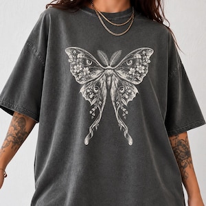 May include: A charcoal gray short-sleeve t-shirt featuring a large, detailed white graphic print of a moth with intricate wing patterns, floral elements, and eye-like markings. The shirt has a relaxed, oversized fit with a crew neckline.