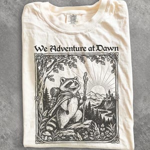 May include: A cream-colored t-shirt featuring a black ink illustration of a raccoon wearing a cape and carrying a backpack and walking stick. The graphic includes the text We Adventure at Dawn, a forest, mountains, and a castle under a rising sun.