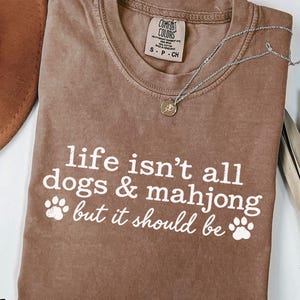 May include: A brown short-sleeved t-shirt with white text that reads: life isn't all dogs & mahjong but it should be. The shirt features two white paw print graphics and a Comfort Colors brand label at the inner neckline. A silver necklace is visible.