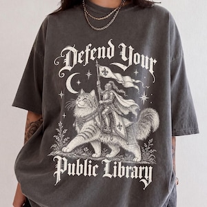 May include: A charcoal grey short-sleeve t-shirt featuring a white graphic print of a knight in armour riding a large cat. The text "Defend Your Public Library" is printed in a gothic font above and below the illustration, which includes a crescent moon, stars, and a flag.
