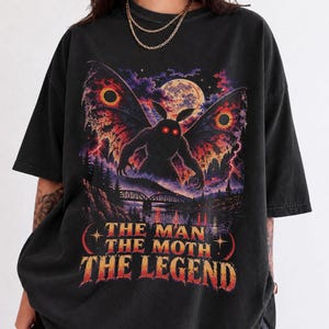 May include: A black short-sleeve t-shirt featuring a graphic print of a winged moth creature with glowing red eyes, a full moon, and a bridge over water. The text reads THE MAN THE MOTH THE LEGEND in a bold, distressed, orange and yellow font.