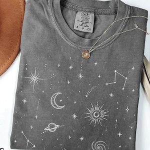 Könnte beinhalten: Ein anthrazitfarbenes Kurzarm-T-Shirt mit einem weißen Himmelsdruck aus Sternen, einer Mondsichel, einem Planeten mit Ringen, einer Sonne und einer Spiralgalaxie. Das Shirt hat einen Rundhalsausschnitt und ein Comfort Colors Etikett am Innenkragen.