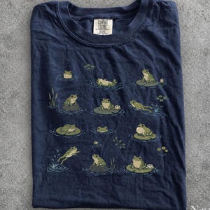 May include: A navy blue t-shirt featuring a graphic print of various green frogs in different poses, including sitting on lily pads, swimming, and resting on rocks. The shirt has a crew neckline and a Comfort Colors brand label at the inner collar.
