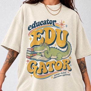 Puede incluir: Una camiseta beige de manga corta con un gráfico de un caimán verde con gafas, leyendo un libro junto a una pila de libros. El texto dice "educator EDU GATOR" y "swamp-tested. classroom approved." con detalles de estrellas coloridas.