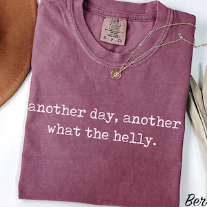 以下が含まれることがあります： ベリー色の半袖Tシャツで、クルーネックと白いプリント文字「another day, another what the helly.」が特徴です。襟の内側にはComfort Colorsのブランドラベルがあり、ゴールドトーンのネックレスが添えられています。
