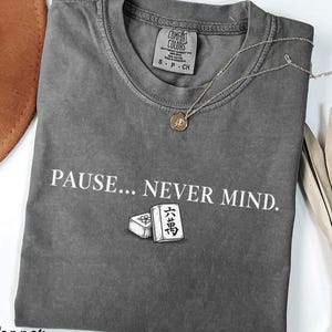 May include: A charcoal gray short-sleeve t-shirt with a crew neckline. The shirt features the white text "PAUSE... NEVER MIND." above a graphic of two mahjong tiles. A thin gold-tone necklace with a small circular pendant rests on the collar.