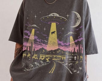 May include: A charcoal grey short-sleeve t-shirt featuring a graphic print of a desert landscape at night with three UFOs beaming light down onto a cow. The scene includes cacti, mountains, a crescent moon, and a road sign reading ROSWELL NM.
