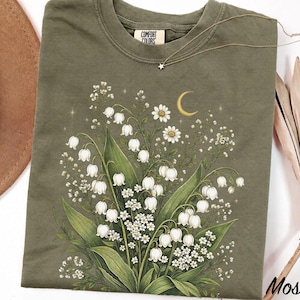 May include: An olive green crew neck t-shirt featuring a graphic print of white lily of the valley flowers, small white daisies, and a yellow crescent moon. The shirt has a Comfort Colors brand label at the inner neckline and short sleeves.