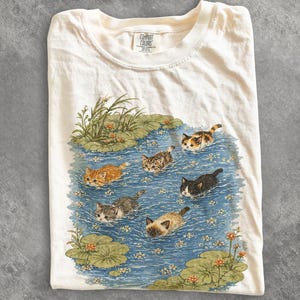 May include: A cream-colored t-shirt featuring a graphic print of six kittens swimming in a blue pond with green lily pads and small orange flowers. The inner neck label displays the text Comfort Colors.