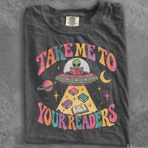 May include: A charcoal gray t-shirt featuring a graphic of an alien in a flying saucer reading a book. The text reads "TAKE ME TO YOUR READERS" in colorful, wavy letters. The design includes a planet, stars, a crescent moon, and books.