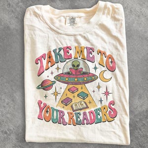 May include: A cream-coloured t-shirt featuring a graphic of a green alien in a flying saucer reading a book. The text reads "TAKE ME TO YOUR READERS" in colourful, curved lettering. The design includes planets, stars, and floating books.