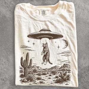 Könnte beinhalten: Ein cremefarbenes T-Shirt mit einem dunkelbraunen Grafikdruck einer Katze, die von einem UFO in einer Wüstenlandschaft mit Kaktus, Bergen, Sternen und einer Mondsichel entführt wird. Das innere Nackenetikett zeigt den Text Comfort Colors.