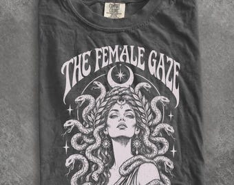 May include: A charcoal gray t-shirt featuring a white graphic print of a person with snakes for hair, reminiscent of Medusa. Above the figure, the text reads THE FEMALE GAZE in a curved font. A crescent moon and stars surround the central illustration.
