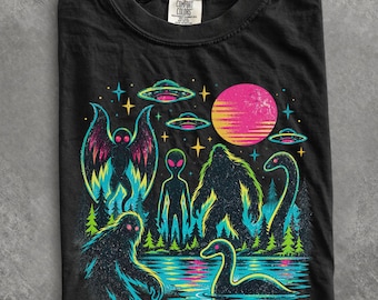 May include: A black t-shirt featuring a vibrant graphic print of cryptids and extraterrestrials, including a moth-like creature, an alien, a sasquatch, and a sea serpent, set against a forest landscape with UFOs, stars, and a large pink sun.