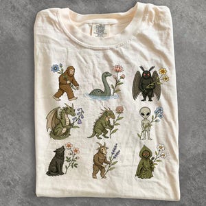 Può includere: Una t-shirt color crema con un'etichetta Comfort Colors sul collo. La parte anteriore presenta una griglia di nove illustrazioni di creature mitiche, tra cui Bigfoot, il mostro di Loch Ness e un uomo falena, ognuna con un fiore selvatico.
