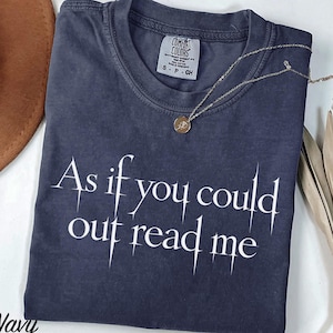 May include: A navy blue short-sleeve t-shirt with a crew neckline and a white graphic print that reads As if you could out read me. The shirt features a Comfort Colors brand label on the inside collar indicating a size small and 100% cotton material.