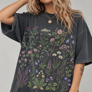 May include: A charcoal gray short-sleeve t-shirt featuring a detailed botanical print of various wildflowers in shades of pink, purple, blue, white, and green. The shirt has a relaxed fit with a crew neckline and a faded, washed-out fabric texture.