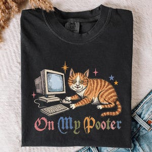 Peut inclure: Un t-shirt noir à col rond avec un motif représentant un chat tigré orange assis à côté d'un ordinateur et d'un clavier vintage. Le texte "On My Pooter" est imprimé en dessous dans une typographie colorée et stylisée, entouré d'étoiles.