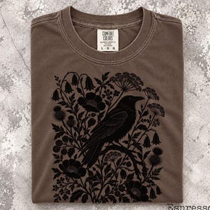 May include: A brown crew neck t-shirt featuring a black graphic print of a crow perched on a branch surrounded by various wildflowers and thistles. The inner neck label displays the text Comfort Colors and the size L.