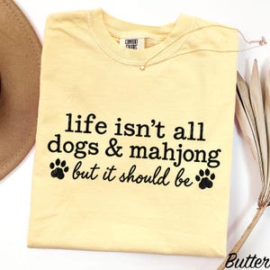 May include: A pale yellow short-sleeved t-shirt with black text that reads, "life isn't all dogs & mahjong but it should be," accompanied by two black paw print graphics. The shirt features a Comfort Colors brand label at the inner neckline.