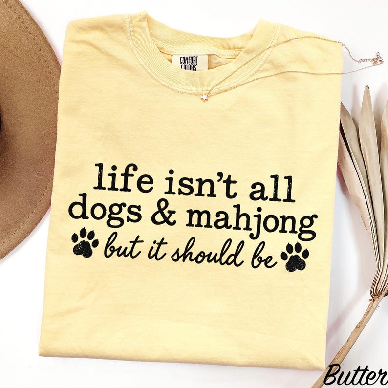 May include: A pale yellow short-sleeved t-shirt with black text that reads, "life isn't all dogs & mahjong but it should be," accompanied by two black paw print graphics. The shirt features a Comfort Colors brand label at the inner neckline.