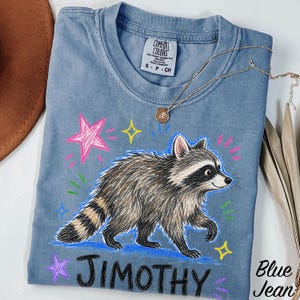 May include: A blue short-sleeved t-shirt featuring a graphic of a raccoon walking, surrounded by colorful hand-drawn style stars and dashes. The text JIMOTHY is printed in black below the raccoon. The shirt has a Comfort Colors brand label.